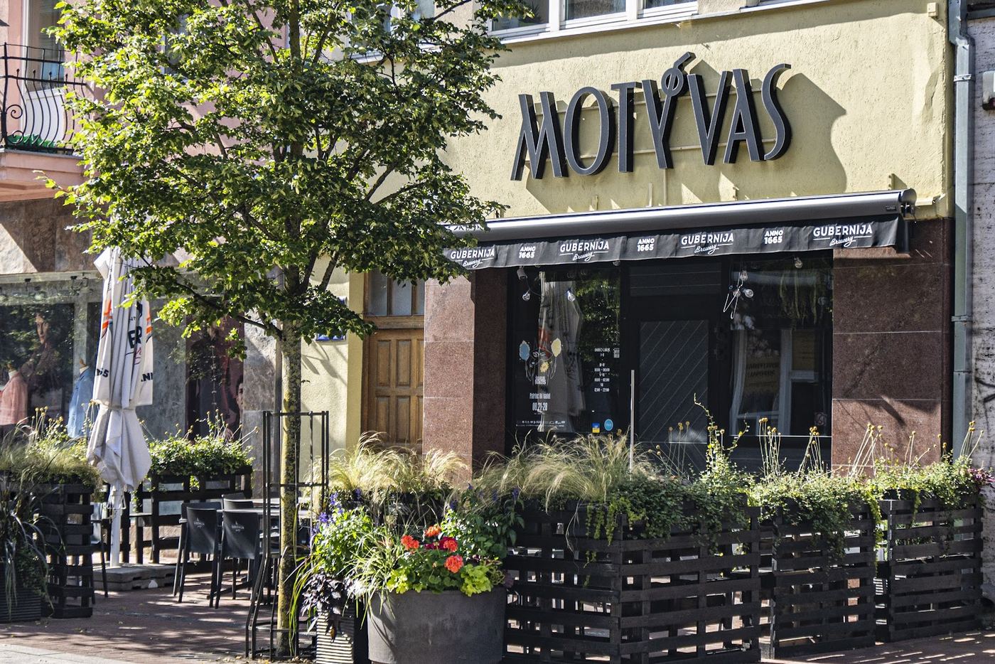 Motyvo fasadas su užrašu MOTÝVAS ir terasa su gėlėmis Vilniaus gatvėje
